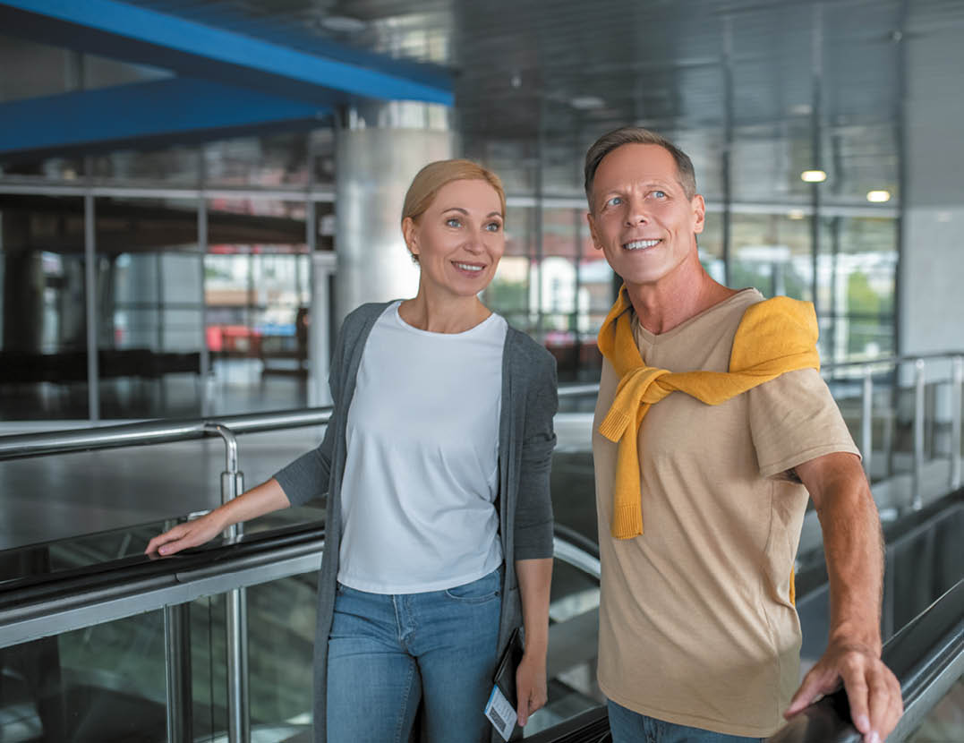Smiling female tourist with travel documents and her happy companion with the luggage stepping off the moving staircase